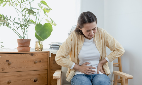 A girl is sitting on sofa and she is holding her stomach with both hands as she is having digestive issues. She is concerned with a question about how to improve digestion.